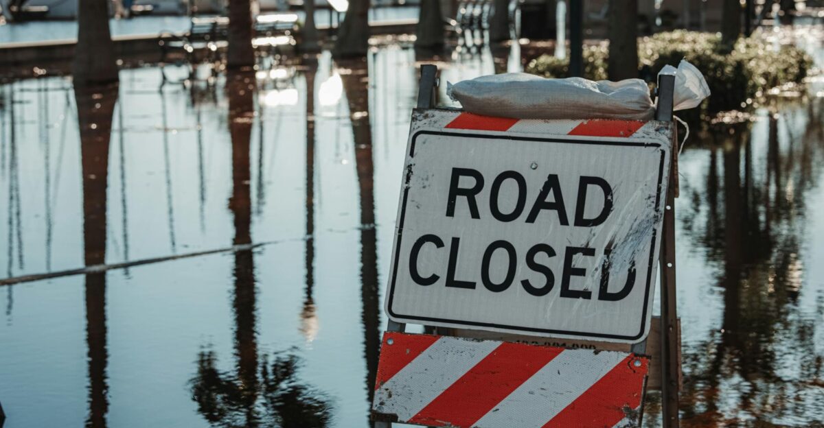 Road closed sign amidst flooded street reflecting calm water and trees