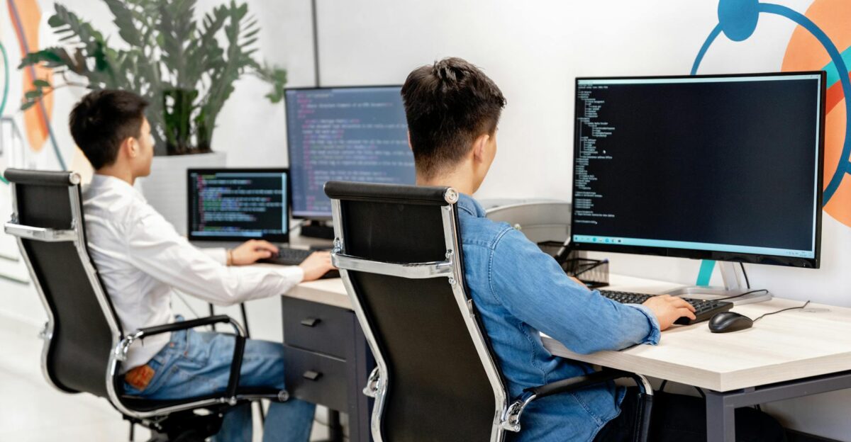 Two male developers at desks programming in a modern office workspace with large monitors